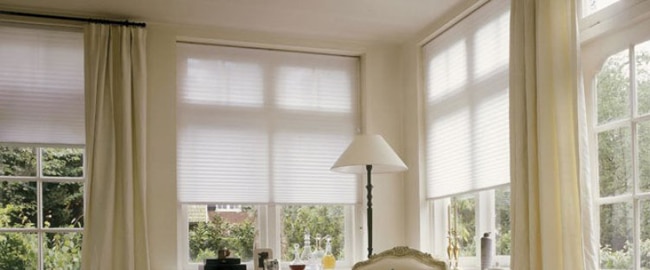 Honeycomb Honeycomb Blinds 19 Bright room featuring honeycomb blinds on multiple windows, paired with light-colored curtains. A lamp and decorative items are visible.